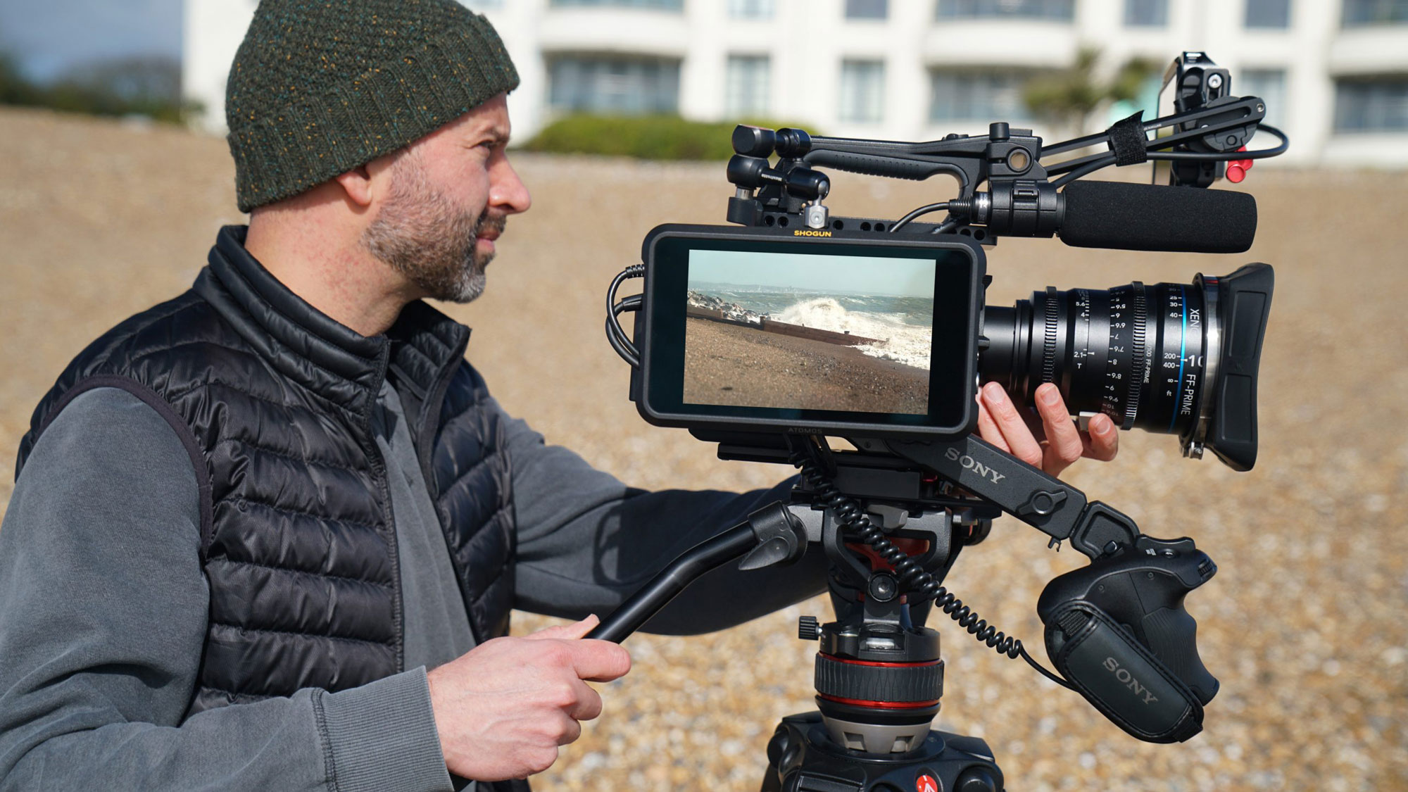 a man shooting beach footage on his video camera. a live feed of the ocean plays on the external camera screen, which is mounted on the side of the video camera