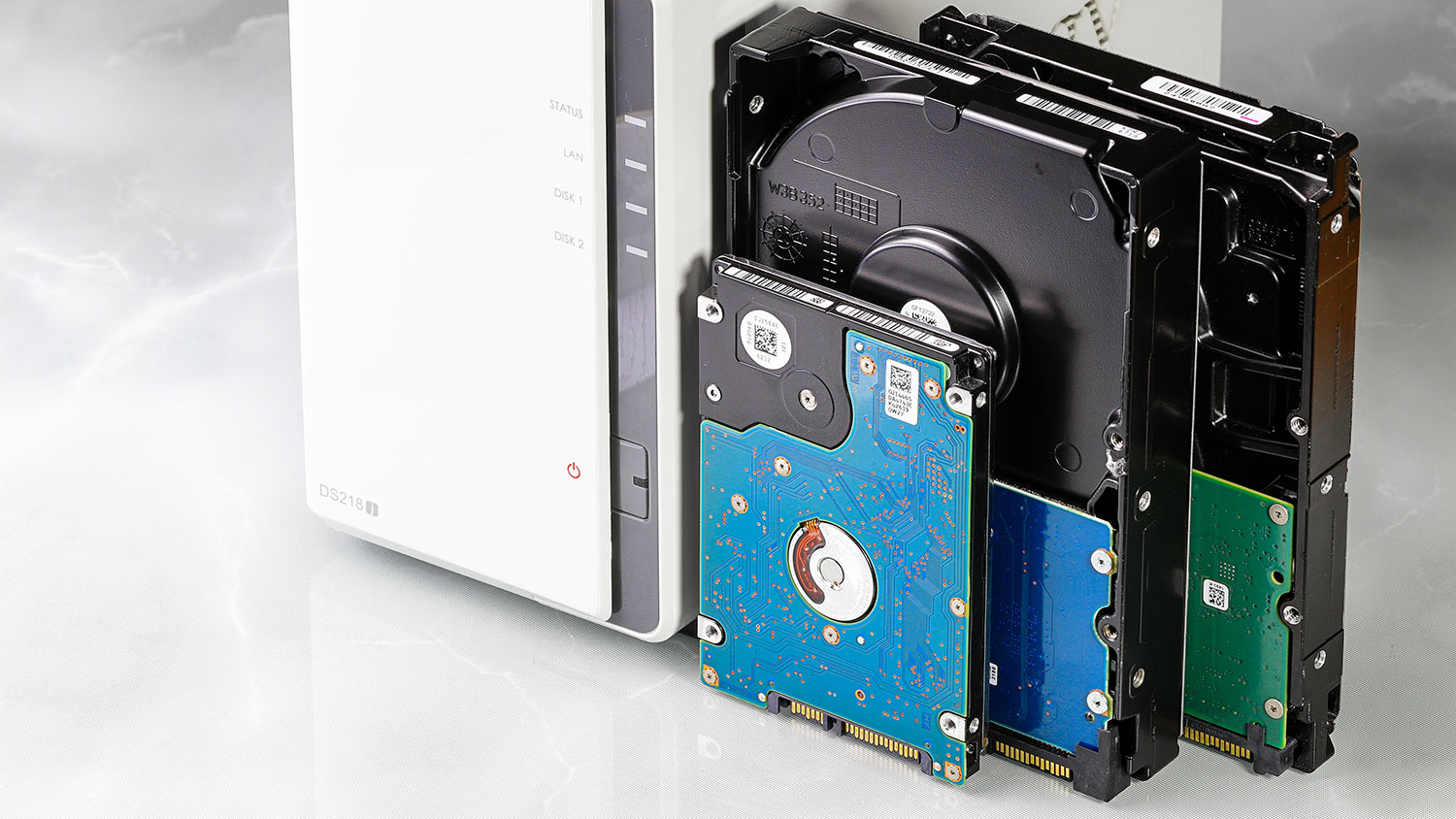 A 2.5-inch hard drive and two 3.5-inch hard drives with exposed circuit boards next to a white NAS storage enclosure on a light surface.